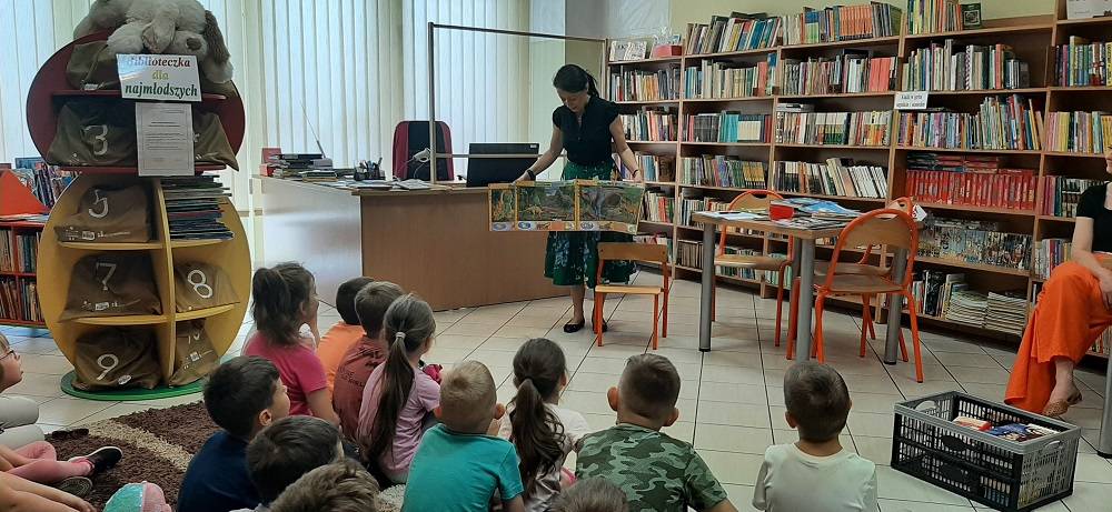 zdjęcie przedstawia dzieci przedszkolne uczestniczące w lekcji bibliotecznej, bibliotekarka pokazuje rozkładaną książkę