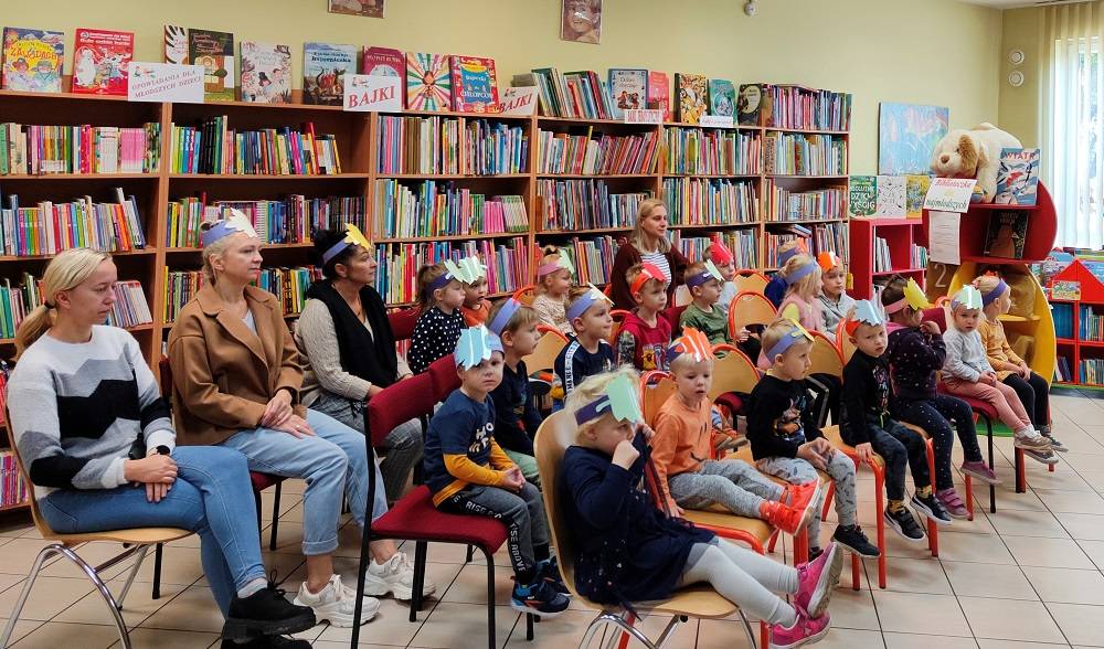 Grupa przedszkolaków siedząc na krzesłach słucha bibliotekarki