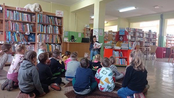 Dzieci siedzać na dywanie słuchają czytanej przez bibliotekarkę książki.
